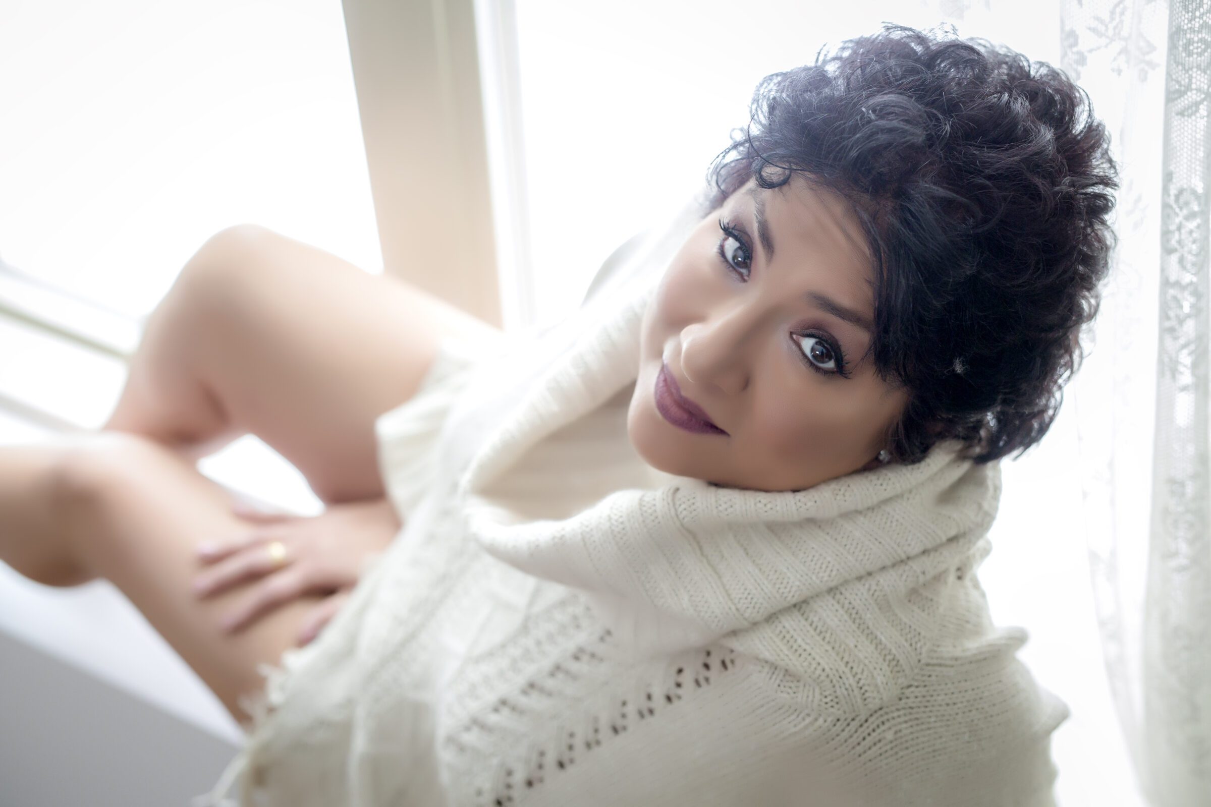 mature woman wearing a cream turtleneck, sitting on a window ledge while posing for a boudoir photographer